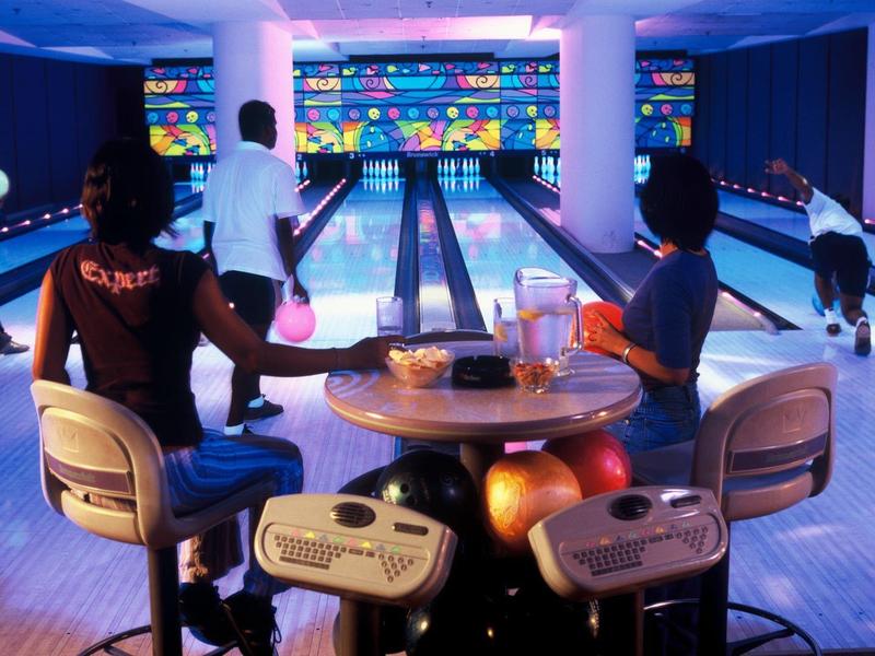 Zwei Personen sitzen an einem Tisch in einer modernen Bowlingbahn mit beleuchtetem Hintergrund.