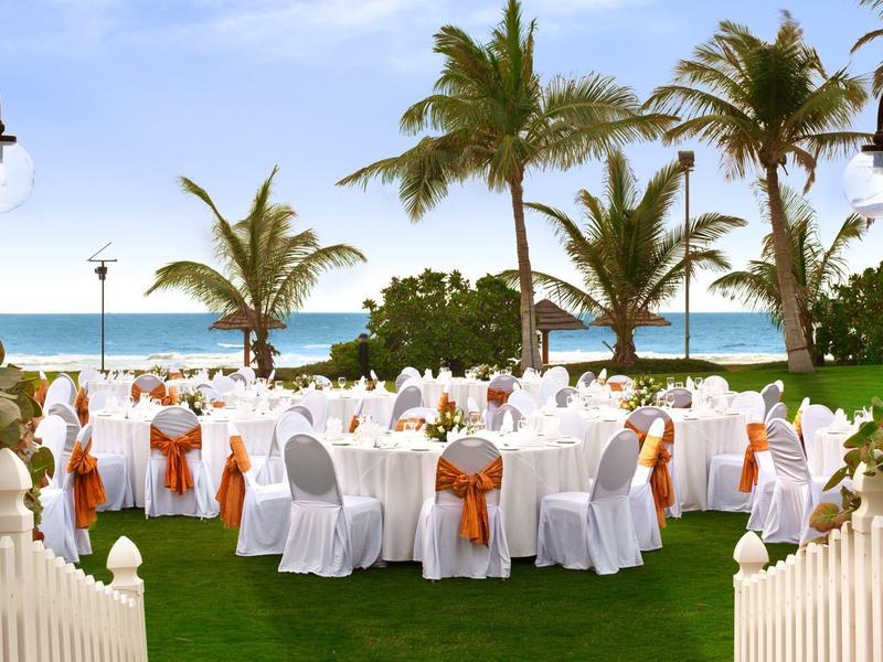 Elegantes Freiluft-Bankett mit weißen Tischen und Stühlen, orangefarbene Schleifen, Palmen am Strand.