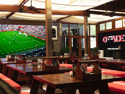 Příjemná restaurace s dřevěným nábytkem a velkou televizí ukazující fotbalový zápas.