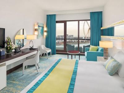 Helles Hotelzimmer mit großem Bett, Schreibtisch, Sitzecke und Balkon mit Hafenblick.