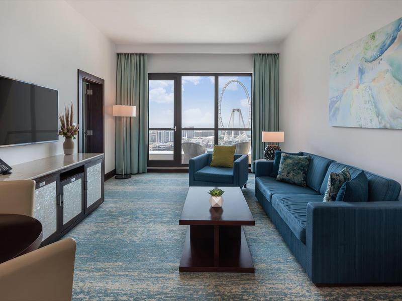 Chambre d'hôtel lumineuse avec canapé bleu, tapis, grande fenêtre et vue sur la ville.