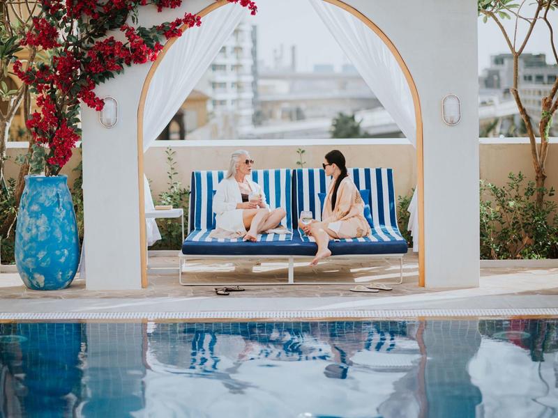 Deux femmes sont assises sous une arche sur un canapé rayé bleu près de la piscine.
