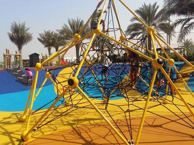 Buntes Klettergerüst aus Seilen auf gelbem Boden in sonnigem Outdoor-Spielplatz mit Palmen.
