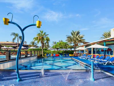 Freizeitpool mit Wasserspielen, blauer Pflasterung, Liegestühlen und Palmen bei klarem Himmel.