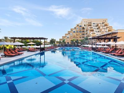 Großer blauer Pool mit Liegestühlen und Sonnenschirmen vor einem terrassenförmigen Hotelgebäude.