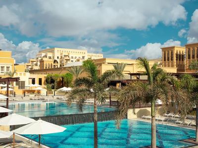Großer Hotelpool mit Palmen, Sonnenschirmen und sandfarbenen Gebäuden unter blauem Himmel.