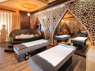 Luxuriöser Spa-Raum mit zwei Massagebänken, runder Badewanne und ornaten Wandelementen in warmem Licht.