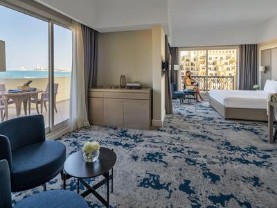 Helles Schlafzimmer mit Meerblick, blauer Teppich, zwei Sesseln und Balkon mit Tisch und Stühlen.