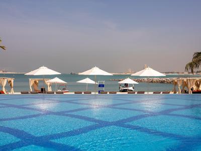 Großer blauer Pool mit weißen Sonnenschirmen am Meer bei klarem Himmel.