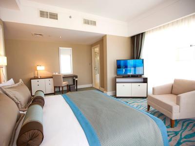 Modernes Hotelzimmer mit großem Bett, Schreibtisch, Fernseher und gemütlichem Sessel am Fenster.