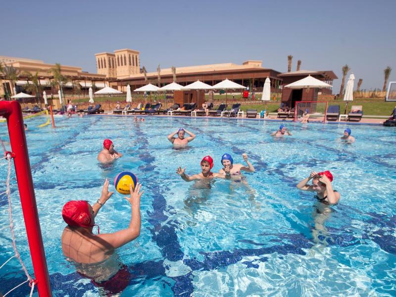 Menschen spielen Wasserball in einem Außenpool bei sonnigem Wetter neben einem Hotel.