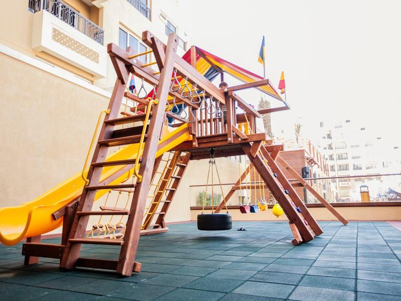 Ein bunter hölzerner Kinderspielplatz mit Rutsche und Schaukeln auf einem gepflasterten Bereich.