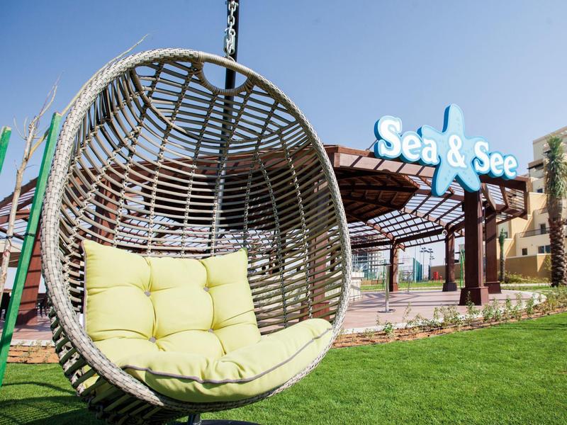 Hängender Korbsessel mit Kissen vor einem Gebäude mit dem Schild ‚Sea & See‘ bei klarem Himmel.