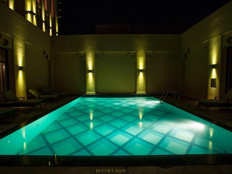 Beleuchteter Swimmingpool in einem Innenhof bei Nacht mit Sitzgelegenheiten an den Seiten.