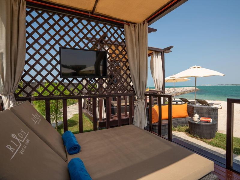 Gemütliche Strandlounge mit Bett, Fernseher und Blick auf das Meer bei sonnigem Wetter.