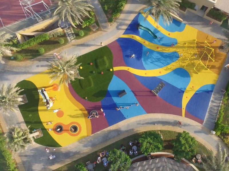 Bunter Spielplatz mit Klettergerüst und Schaukeln umgeben von Palmen und Wegen.