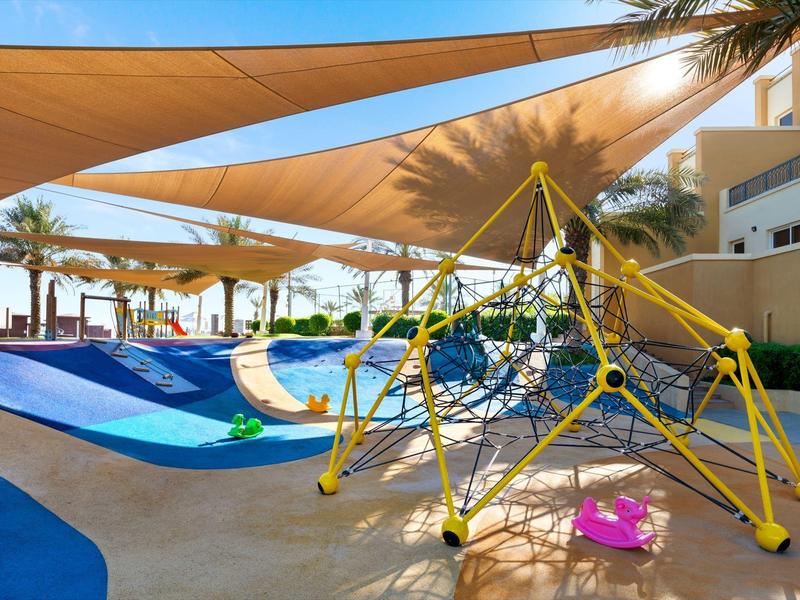 Sonnengeschützter Spielplatz mit Klettergerüst und Rutschbahn vor einem Hotelgebäude.