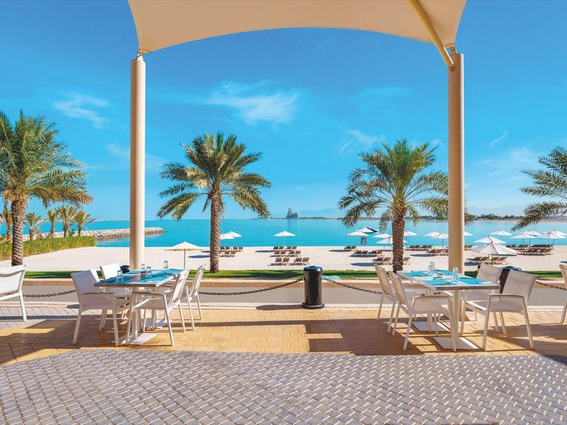 Blick von einer überdachten Terrasse mit Tischen und Stühlen auf einen Strand mit Palmen und blauem Meer.