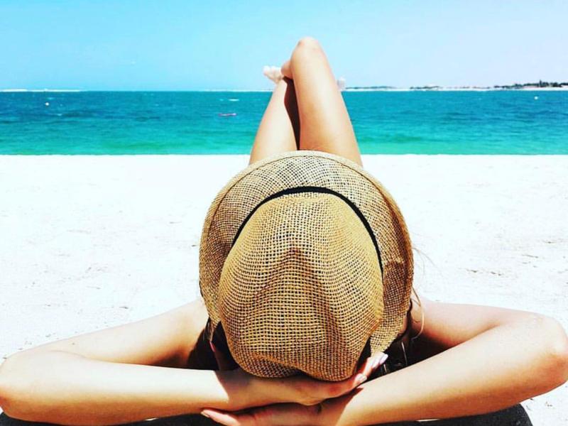 Person liegt am weißen Sandstrand, trägt Strohhut, Blick auf türkisfarbenes Meer und blauen Himmel.