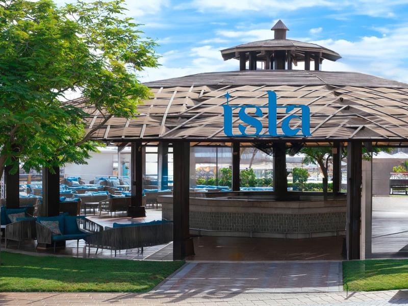 Offener Pavillon mit Holzdach und Bar, umgeben von Bäumen und Sitzbereichen.