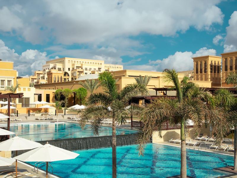 Luxuriöser Hotelpool mit Palmen, Sonnenschirmen und Gebäuden im Hintergrund unter blauem Himmel.