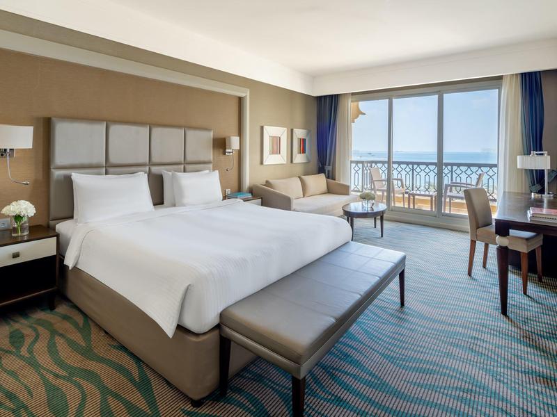 Hell eingerichtetes Hotelzimmer mit King-Size-Bett, Sofa und Meerblick vom Balkon.