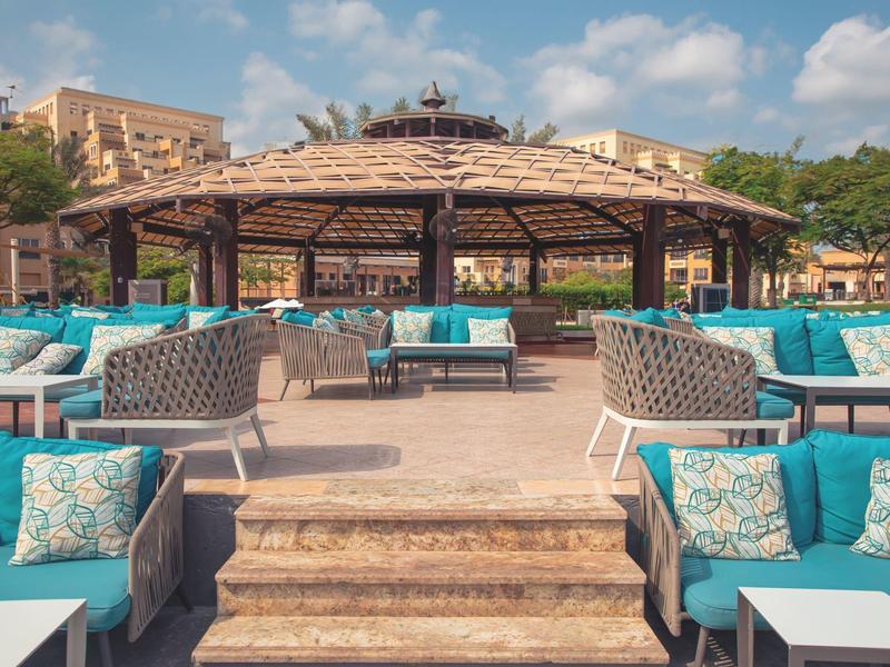 Außenbereich eines Hotels mit Sitzbereichen und einem überdachten Pavillon bei sonnigem Wetter.