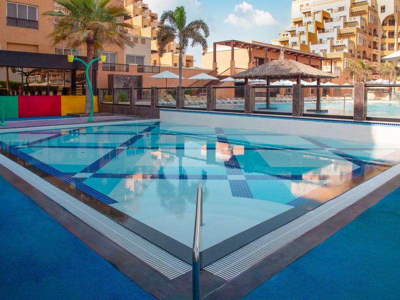 Kleiner Pool mit Wasserspielen und Palmen in einem Hotelkomplex bei Sonnenschein.