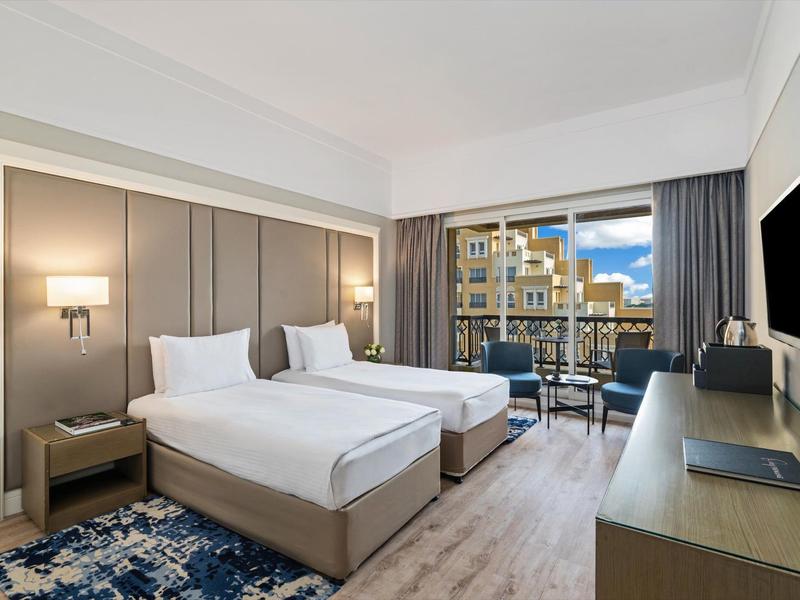 Modernes Hotelzimmer mit zwei Einzelbetten, Schreibtisch und Balkon mit Blick auf Gebäude.