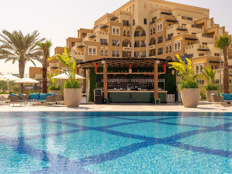 Blick auf einen Pool mit blauem Wasser und einer Bar davor, umgeben von einer Hotelanlage und Palmen.