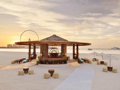 Strandbar mit Holzbau, Sitzplätzen und Riesenrad im Hintergrund bei Sonnenuntergang.