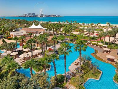 Großer Hotelpool umgeben von Palmen, Sonnenschirmen, Grünflächen und Blick auf das blaue Meer.