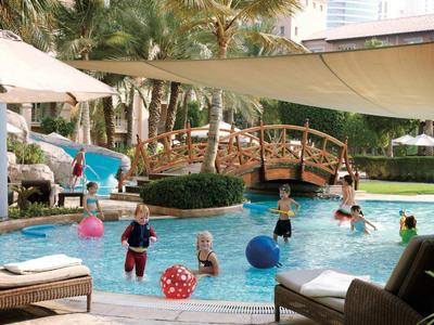 Kinder spielen mit bunten Bällen im Pool, daneben Holzbrücke und Sonnensegel, grüne Pflanzen und Stadtblick.