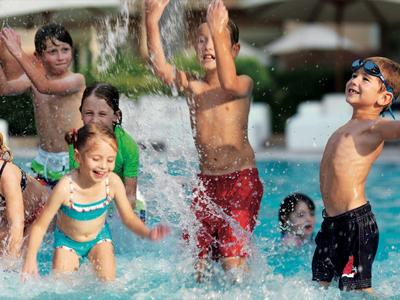 Kinder verschiedener Altersgruppen spielen fröhlich und planschen im Schwimmbecken bei sonnigem Wetter.