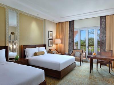 Helles Hotelzimmer mit zwei Betten, Schreibtisch, Sesseln und großen Fenstern mit Vorhängen.