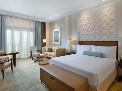 Modern eingerichtetes Hotelzimmer mit großem Bett, Sesseln, Schreibtisch und Balkon im Hintergrund.