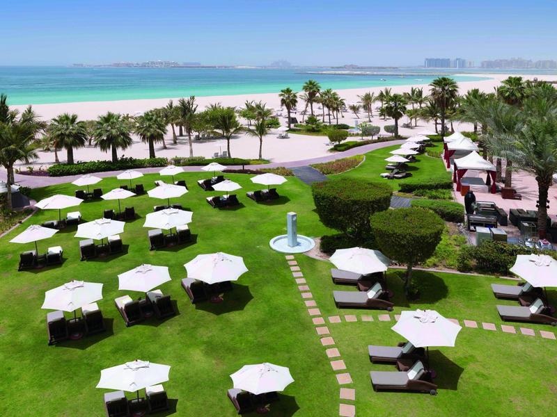 Jardín verde del hotel con sombrillas blancas junto a una playa con vista al mar y palmeras.
