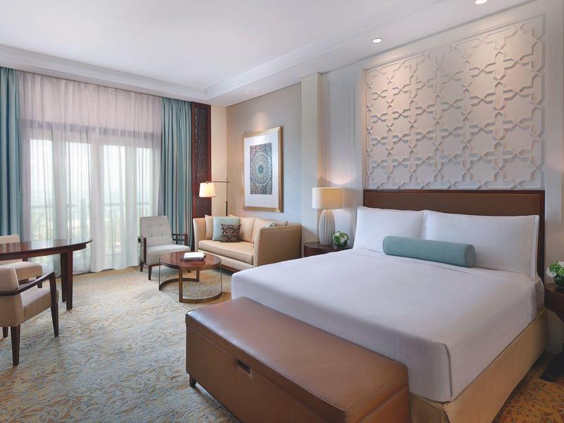 Habitación de hotel elegante con cama grande, área de estar, escritorio y grandes ventanas con cortinas.