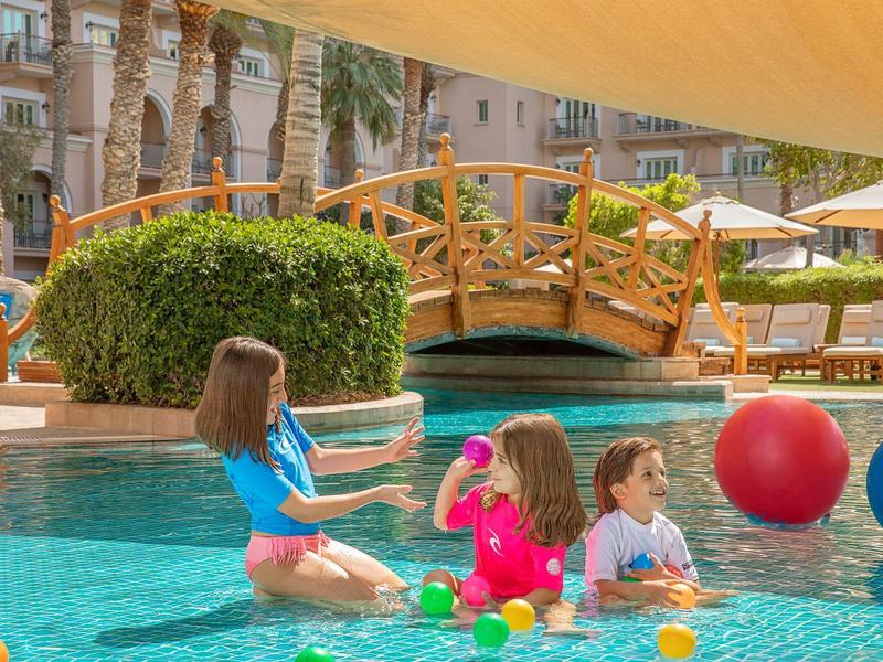 Niños juegan con pelotas de colores en la piscina junto a un puente de madera y palmeras.