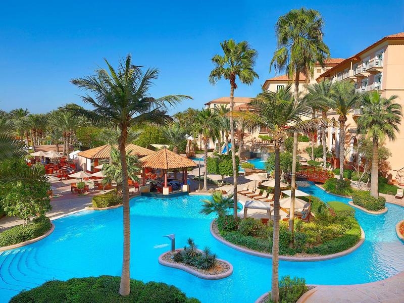 Gran piscina del hotel con palmeras alrededor, tumbonas y cielo azul claro.