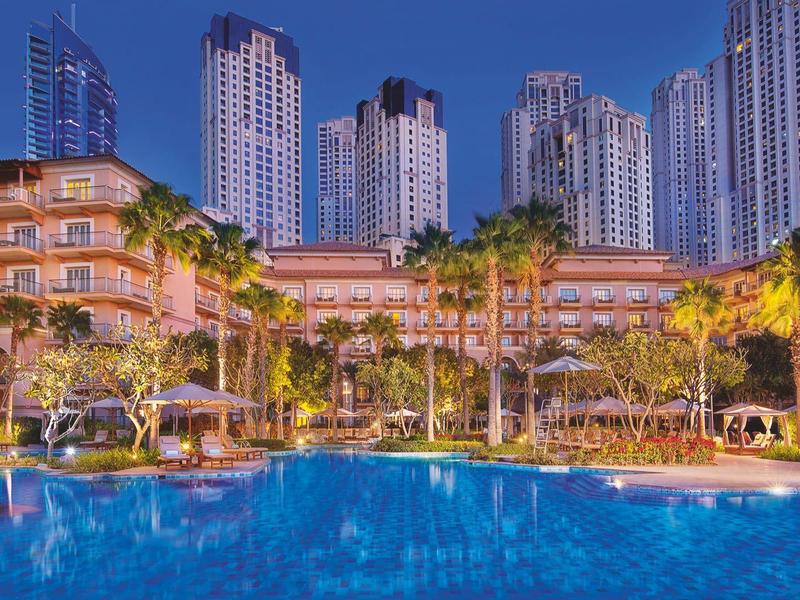 Hotel iluminado con piscina grande y rascacielos al fondo al anochecer.