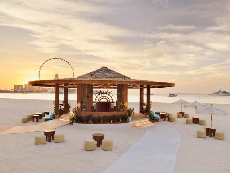 Chiringuito de playa abierto con taburetes y una noria al fondo al atardecer.