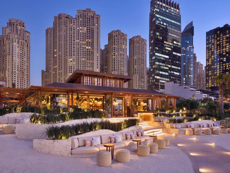 Complejo hotelero moderno con área exterior iluminada y rascacielos al fondo al atardecer