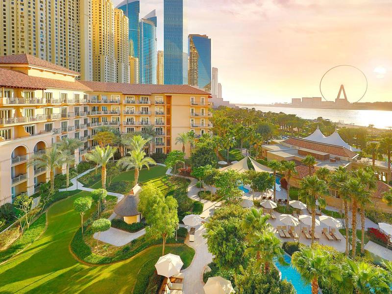Complejo hotelero de lujo con jardín, piscina y vista a una ciudad con rascacielos y noria al atardecer.
