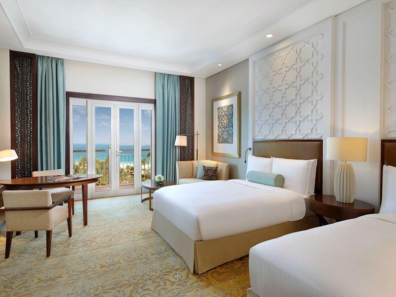 Habitación de hotel elegante con dos camas, escritorio, sillones y vista al mar a través de la ventana.