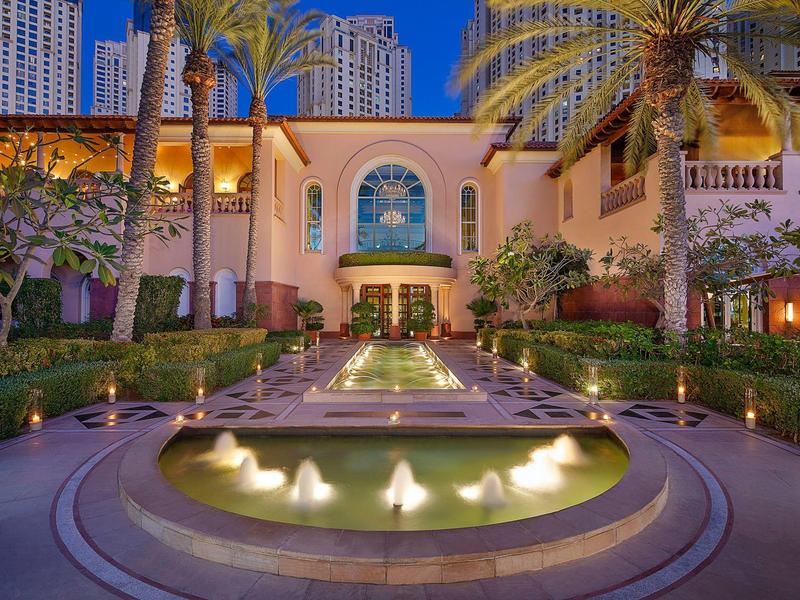 Vista nocturna de un jardín de hotel de lujo con fuente iluminada y palmeras.