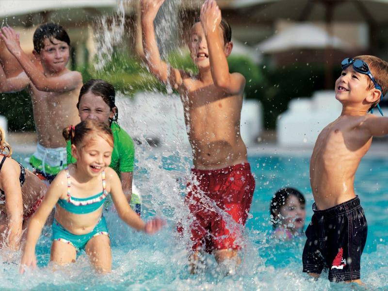 Varios niños juegan y chapotean alegremente en una piscina bajo sombrillas.