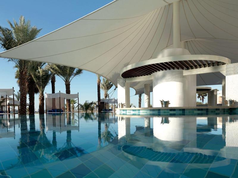 Área de piscina moderna con cubierta, rodeada de palmeras y muebles de descanso bajo cielo despejado.