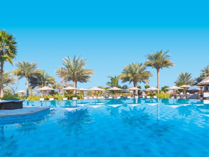 Piscina al aire libre en un resort con palmeras y tumbonas bajo un cielo despejado.