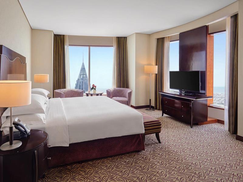 Jasno umeblowany pokój hotelowy z dużym łóżkiem, fotelami i widokiem na miasto.